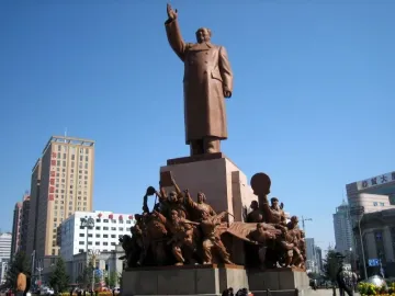 Colossal bronze statue of Mao Zedong with his right arm raised, standing above a multi-tiered base with bronze figures below it.