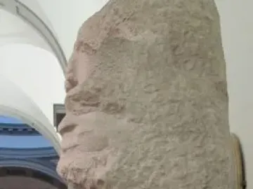 Unfinished marble sculpture of Saint Matthew emerging from a rough stone block, with detailed head, arm, and draped torso.