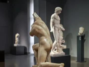 The Subiaco Ephebe sculpture on a dark pedestal in a museum gallery, with other classical statues in the background.