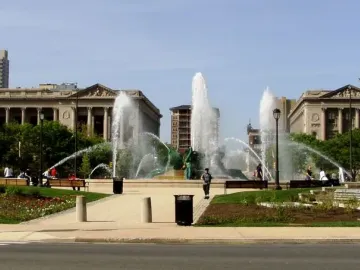 Swann Memorial Fountain in a city plaza, surrounded by paved walkways, trees, and classical stone buildings.
