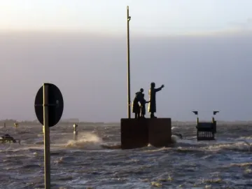 Auswandererdenkmal standing in choppy floodwater under an overcast sky, with a dark circular sign in the foreground.