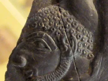Carved stone fragment from the Bull Palette showing a stylized profile head with patterned hair, a detailed eye, and a curly beard split by a vertical crack.