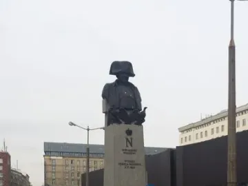 The Emperor Napoleon I Memorial, a dark bust of Napoleon in a bicorne hat on a tall stone plinth in a city square.
