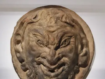 White circular relief sculpture of the Head of a Faun with a grinning face, pointed ears, and curly beard and hair.