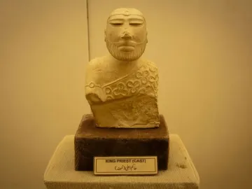 Bust of the Priest King sculpture with a decorative headband and patterned cloak on a dark base