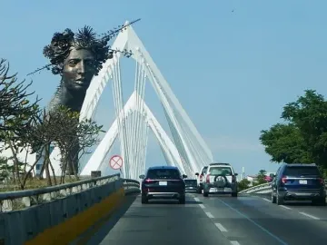 Reminiscencia, a dark bust of a woman crowned with flowers and spiky elements, set before a white cable-stayed bridge.