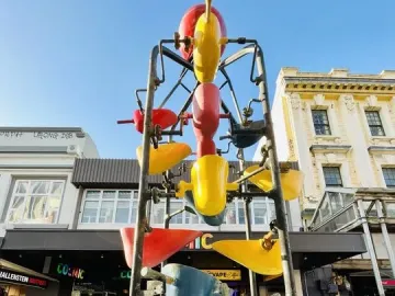 The Bucket Fountain sculpture with colorful buckets in a light blue basin on Cuba Mall, with buildings and shop fronts behind it.