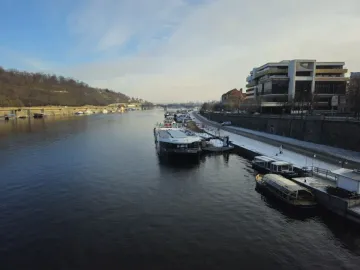 Winter view of the Vltava River with moored boats, a snowy embankment, and a wooded hill across the water; the Prague Metronome is not clearly visible.