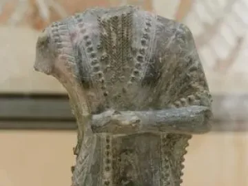 Iddi-Ilum statuette of a headless standing figure with folded arms and a decorated robe on a square base.