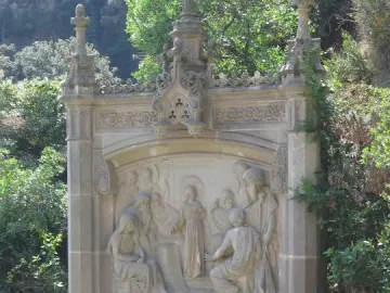 Ornate stone relief on the Monumental Rosary of Montserrat showing several figures gathered around a young person, with decorative architectural details and carved banners.