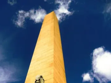 National Women\'s Monument with a tall obelisk and bronze figures of two women on the pedestal against a blue sky.