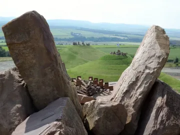 Rugged reddish-brown stones of the Omphalos frame rolling green hills and spiral terracing beyond.