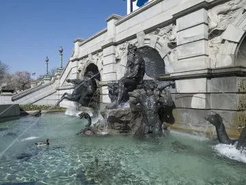 Court of Neptune Fountain with dark metal sea deities and horses rising from a water pool beside an ornate stone building.