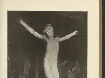 Black and white photograph of a crucifix sculpture showing a nude bearded figure with outstretched arms against a dark background.