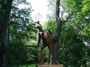 Bronze Greek Warrior Monument of an armored fighter with a shield and raised sword in a forest setting.