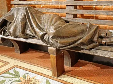 Bronze Homeless Jesus sculpture of a covered figure lying on a slatted bench, with bare feet visible at one end.