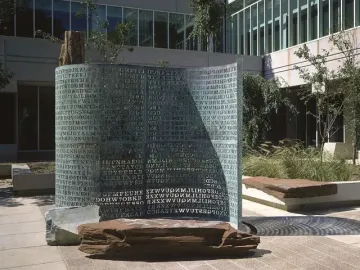 Kryptos bronze sculpture covered with carved capital letters, standing outdoors near stone benches and trees.