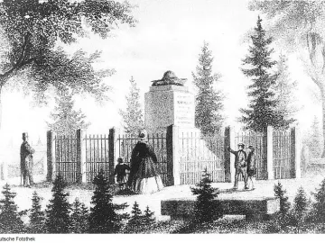 Napoleonstein stone pillar topped with a bicorne hat, enclosed by a fence and surrounded by trees, with several people standing nearby.
