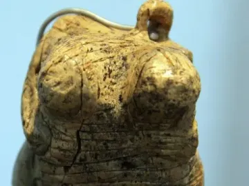 Venus of Hohle Fels figurine with cracked ivory torso, prominent breasts, and a loop at the top on a dark stand.