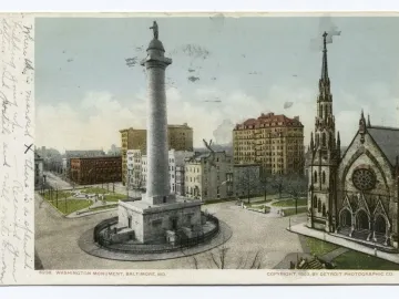 The Washington Monument rises from a public square, with surrounding city buildings and a gothic-style church steeple nearby.