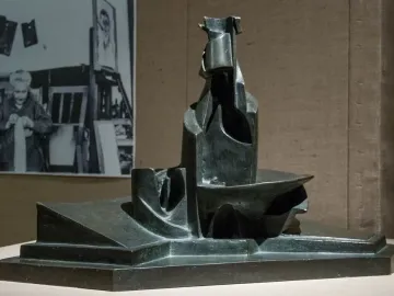 Dark abstract sculpture of Development of a Bottle in Space on a light surface, with a black-and-white studio photograph in the background.