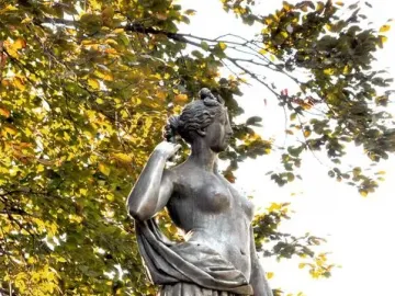 Weathered Diana Fountain statue seen from below against dense green foliage.