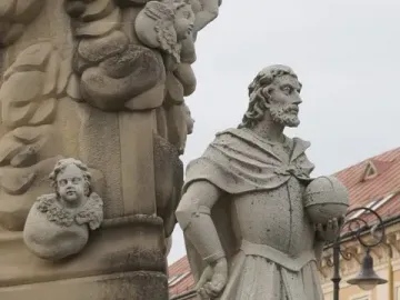 Weathered stone statue beside the Marian Column, with an ornate base decorated with cherub heads.