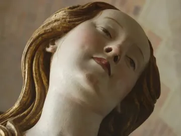 Close-up of Mary Magdalene’s sculpted face with wavy golden hair and red lips.