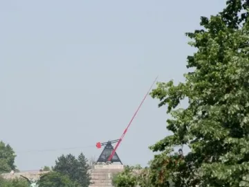 The Prague Metronome, a large red kinetic sculpture, stands on a stone pedestal above a tree-covered hill.