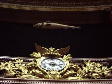 The Sacred Cod hangs horizontally against a dark backdrop above an ornate golden clock with leaf motifs and a winged top.