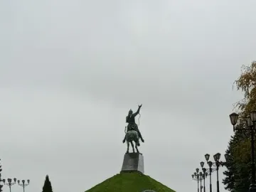 The Salavat Yulaev Monument stands on a grassy mound above a wet walkway, with visitors holding umbrellas on a rainy day.