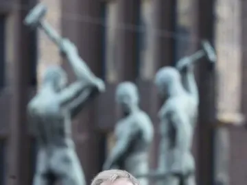 A smiling man in glasses and a suit stands in front of the Three Smiths sculpture.