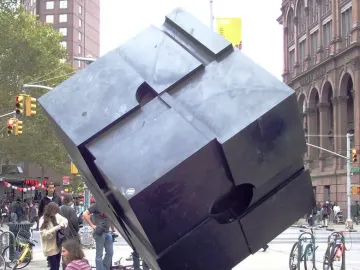 The Alamo, a dark cube sculpture, stands balanced on one corner in an urban plaza with buildings and traffic lights behind it.