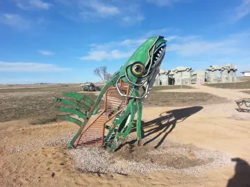 Carhenge-like roadside monument with cars arranged on gray supports behind a large green metal sculpture in the foreground.