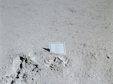 A small white fallen astronaut figure lies beside a plaque on a gray lunar surface.