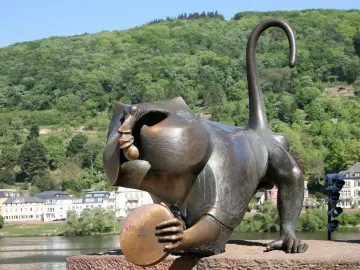 Heidelberg Bridge Monkey sculpture crouching on a stone ledge, holding a round disk with the river and wooded hill behind it.