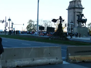 The Bowman statue stands on a stone pedestal beside a monumental archway, overlooking a street with barriers, traffic lights, and people.