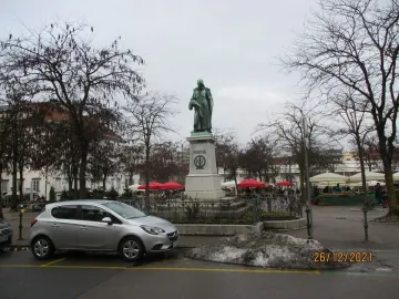Vodnik Monument on a tall stone pedestal in a paved square with bare trees and buildings nearby.