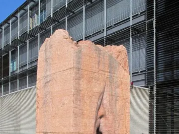 Pi-Chacán, a reddish-orange stone sculpture with a deep vertical crevice, standing on dark gravel beside a modern building.