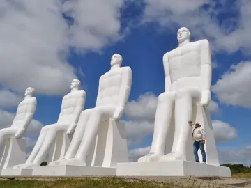 The Man by the Sea sculpture with four massive seated white figures against a blue sky, with a person beside the front statue.