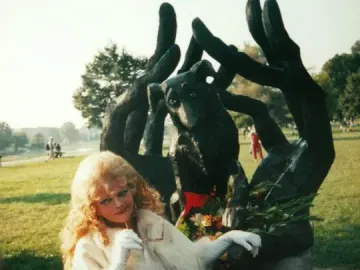 Person standing beside the Monument to Dog Jock, a dark sculpture of a dog held by stylized hands, decorated with a red ribbon and greenery.