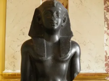 Upper body of a Ptolemaic king statue in dark stone, wearing an Egyptian headcloth and kilt with broken arms.