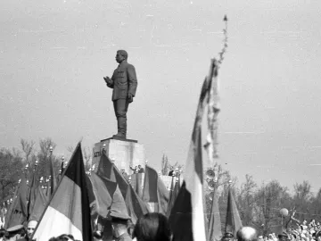 Stalin Monument with a uniformed figure gesturing from a tall pedestal above a crowd holding flags.
