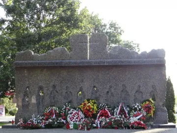Monument to the Victims of the Wola Massacre with a rough memorial wall, carved figures, circular openings, and wreaths at the base.