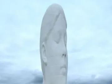 Dream sculpture, a tall white head and neck with closed eyes, standing on a circular base under a cloudy sky.
