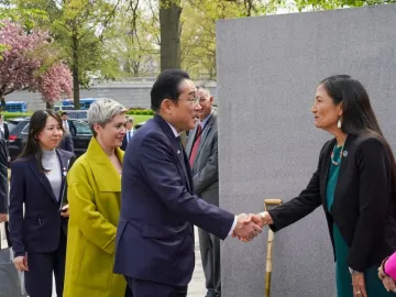 A man and a woman shake hands in front of the Japanese American Memorial, with several people standing nearby and blooming trees in the background.