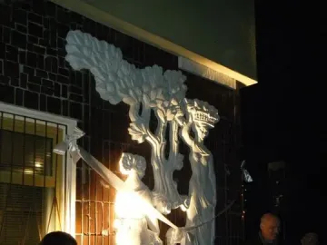 Plon relief sculpture mounted on a dark brick wall, with a woman speaking at microphones in front of it.