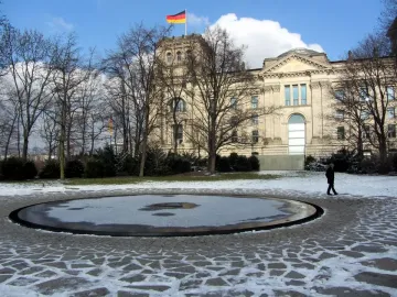 A snow-covered circular basin set in a stone-paved area, with bare trees and a classical-style building in the background.