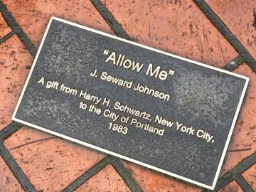 Dark metal plaque for Allow Me mounted on a brick pavement.