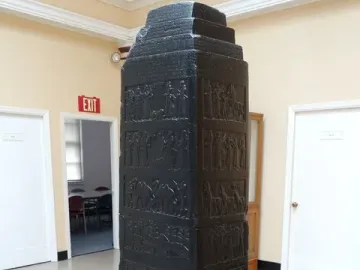 Black Obelisk with stepped top and carved relief bands on a wooden base in a museum interior.